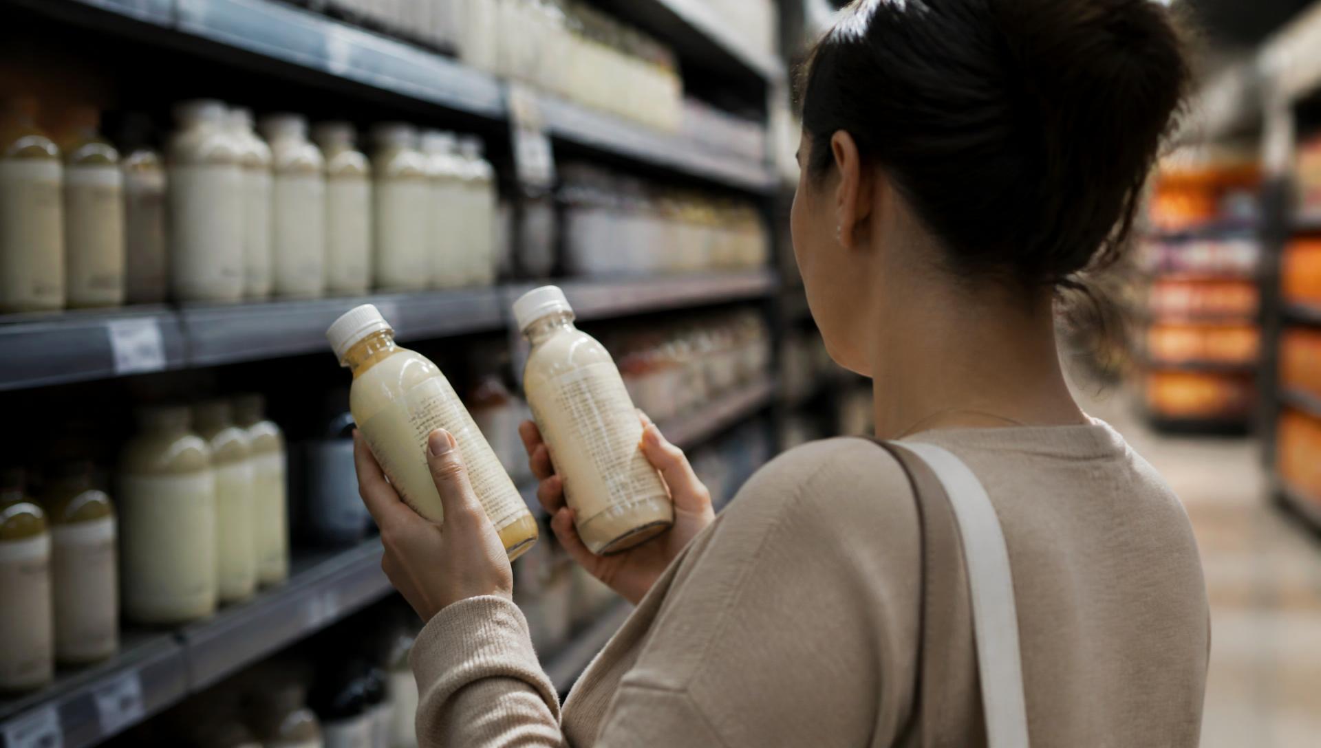 A shopper in a grocery aisle holding two similar bottled drinks, comparing the labels before choosing one.